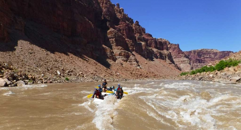 A group of people paddle a raft on whitewater. The river is framed by tall canyon walls.
